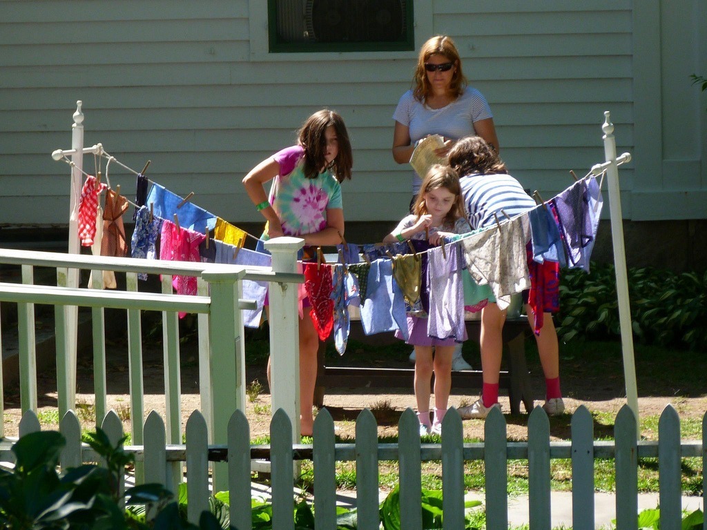 Three children and a person hang colorful laundry on a clothesline outside, beside a white picket fence and house.