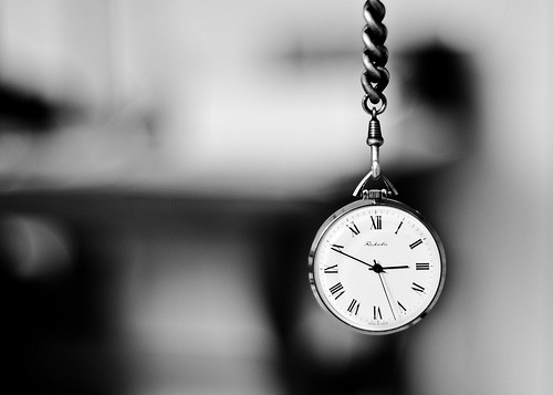 A black-and-white image of a pocket watch with Roman numerals, hanging from a chain against a blurred background, evoking a classic timeless feel.