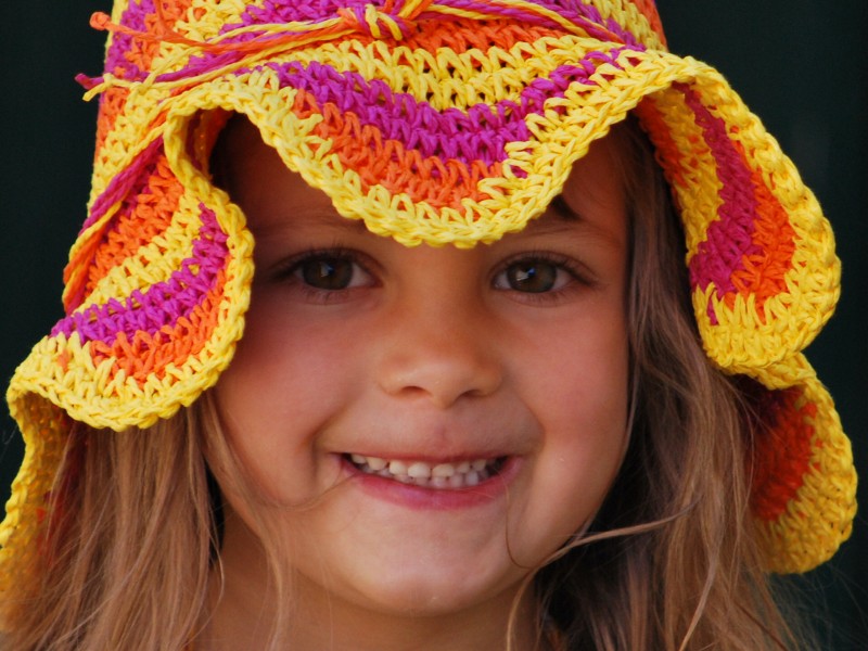 A child smiling, wearing a colorful, striped knit hat with yellow, orange, and purple patterns against a dark background.