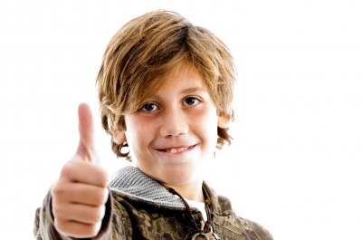 A smiling child with light brown hair gives a thumbs-up against a plain white background, wearing a camouflage-patterned jacket with a hood.