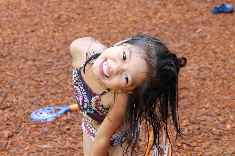 A joyful child in a swimsuit smiles at the camera on a bark-covered ground, with a toy racquet visible nearby.
