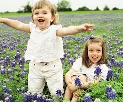 Two children joyfully play in a lush field filled with blooming purple flowers, surrounded by greenery under a clear sky.