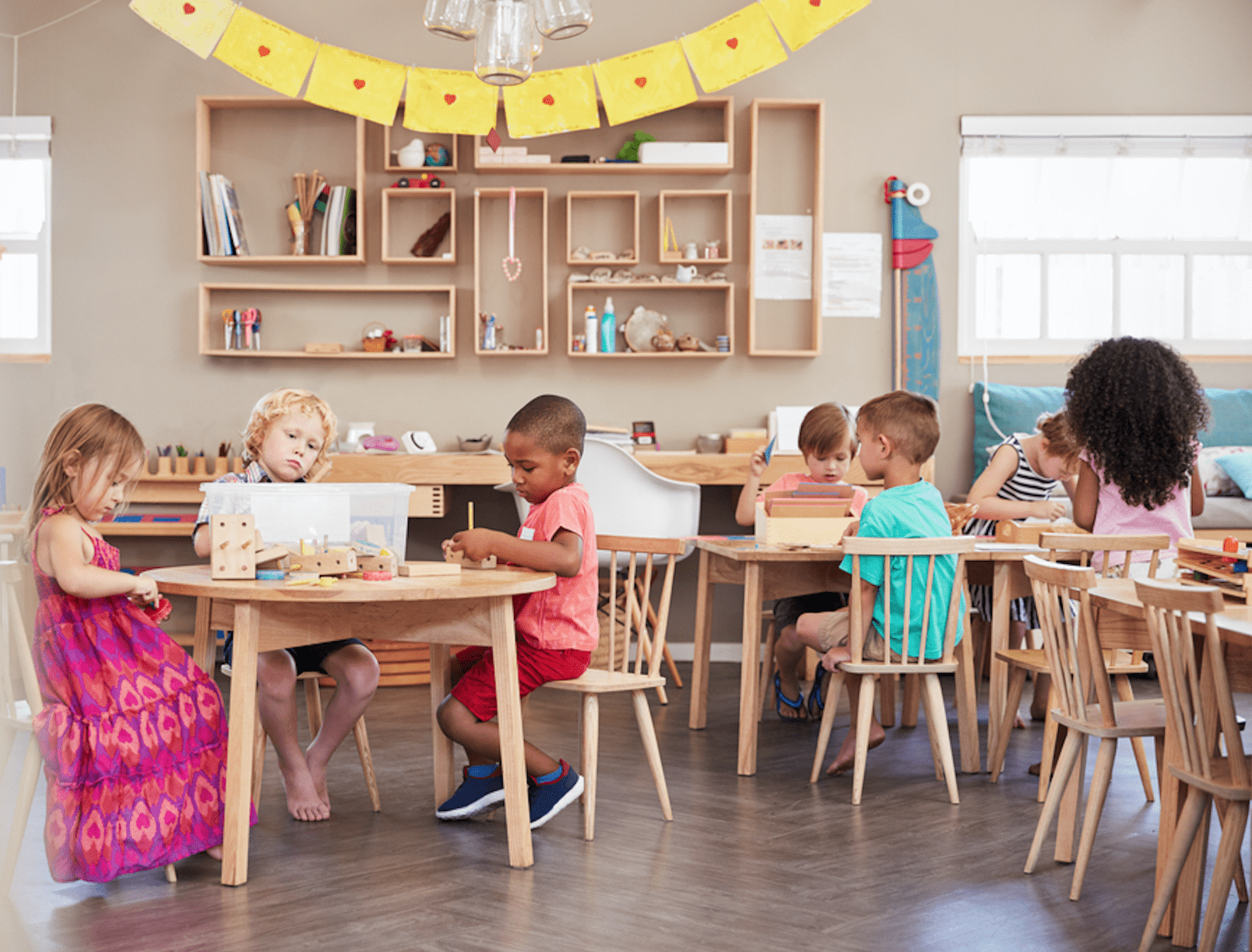 Children are sitting around tables engaged in activities within a well-organized classroom, featuring shelves with educational materials and colorful decorations.