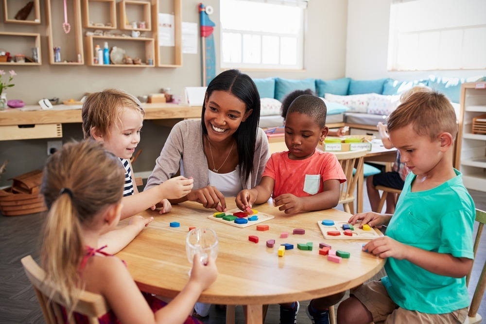 A person and four children play with colorful educational toys around a table in a bright, cheerful classroom setting.