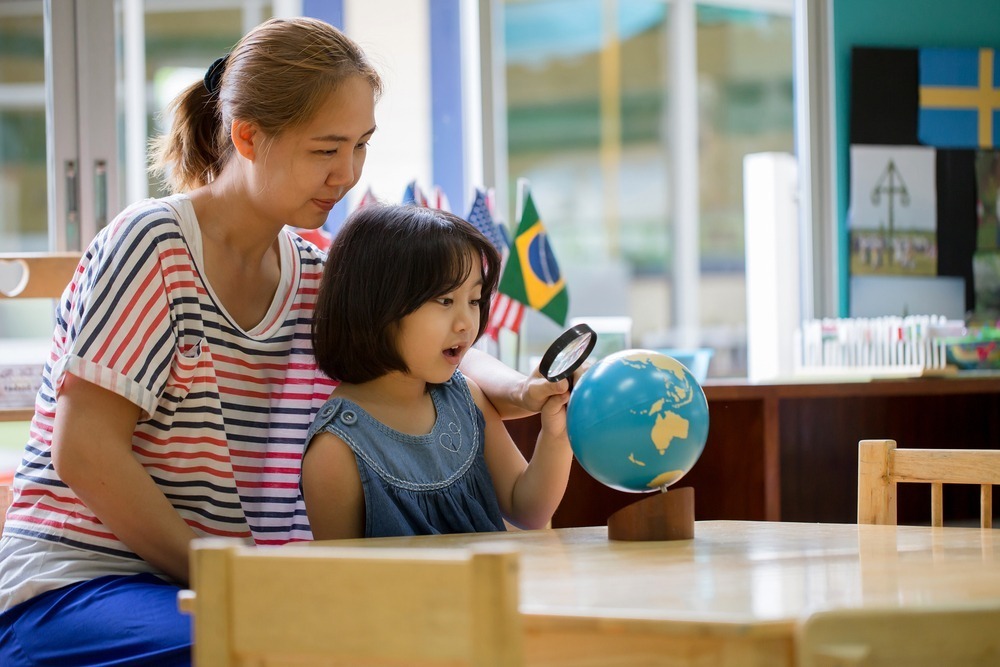 A person and child explore a globe with a magnifying glass in a classroom. Flags are displayed in the background.