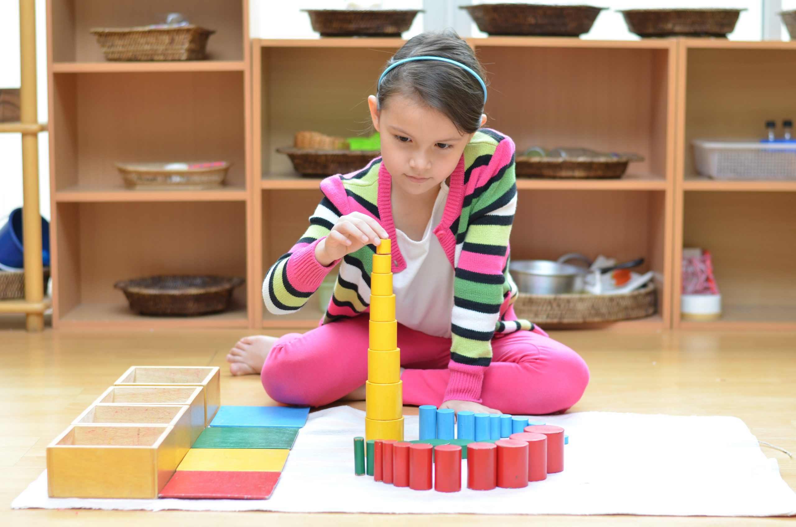 A child is sitting on the floor, focused on stacking colorful wooden cylinders in a room with shelves and baskets.