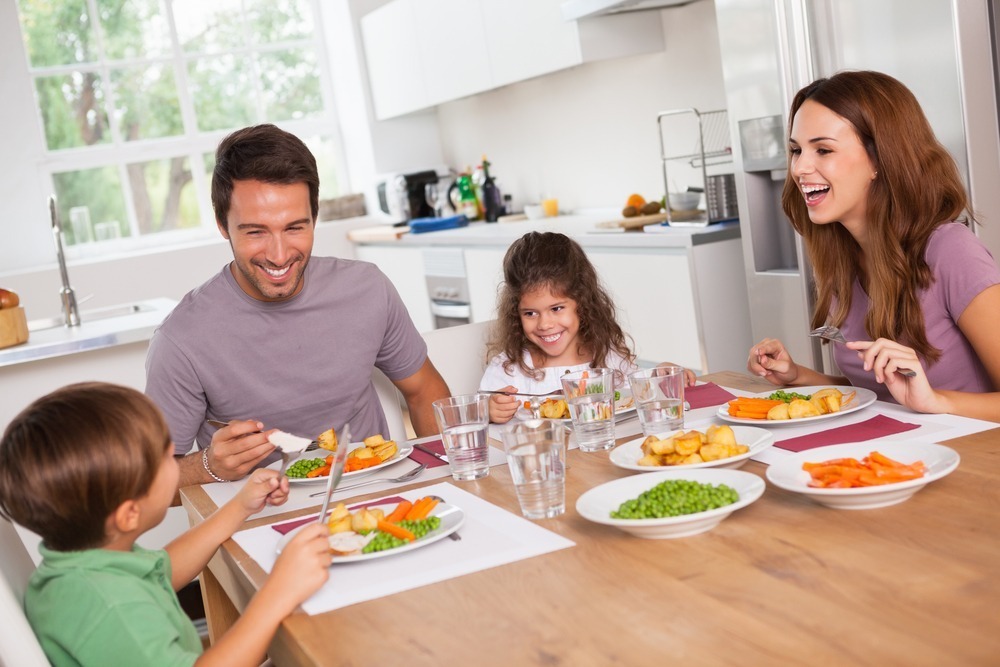 A group of four people, two adults and two children, happily share a meal at a dining table in a modern kitchen.