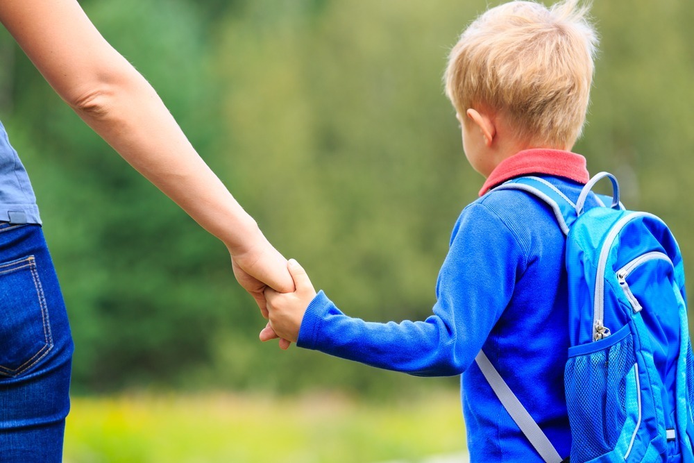 A person holds a child's hand outdoors. The child wears a blue backpack against a blurred, green background, suggesting a natural setting.