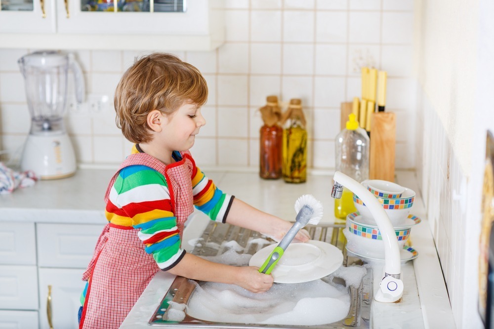 A child in a colorful sweater washes dishes at a kitchen sink, surrounded by bubbles and various kitchen utensils on the counter.
