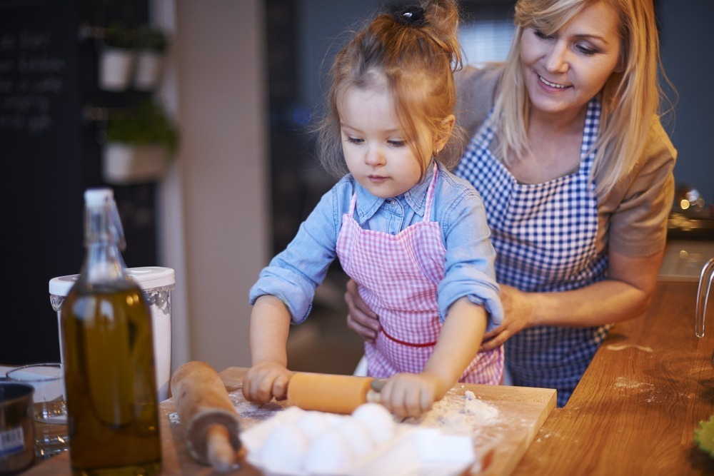 A person helps a child roll dough in a kitchen, surrounded by ingredients like eggs and olive oil. Both wear aprons.