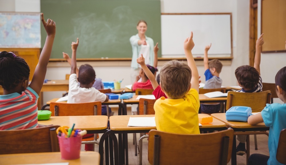 Classroom setting with a person teaching and children raising hands. Desks are arranged in rows, with supplies and a green chalkboard visible.