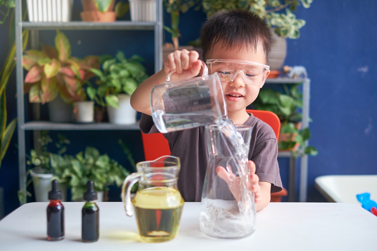 Child wearing safety goggles pours water from pitcher into a glass container. Plants and colorful bottles are in the background.