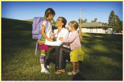 A person kneels beside two children with backpacks in a grassy park, under a sunny sky. Residential buildings are visible in the background.