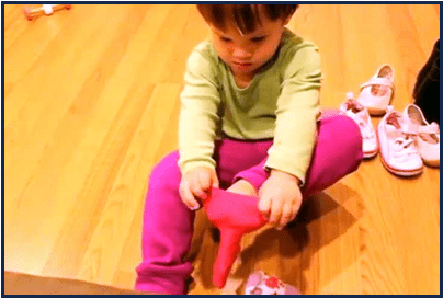 A young child with short hair wears a green top, pink pants, and sits on hardwood floor putting on bright socks near shoes.