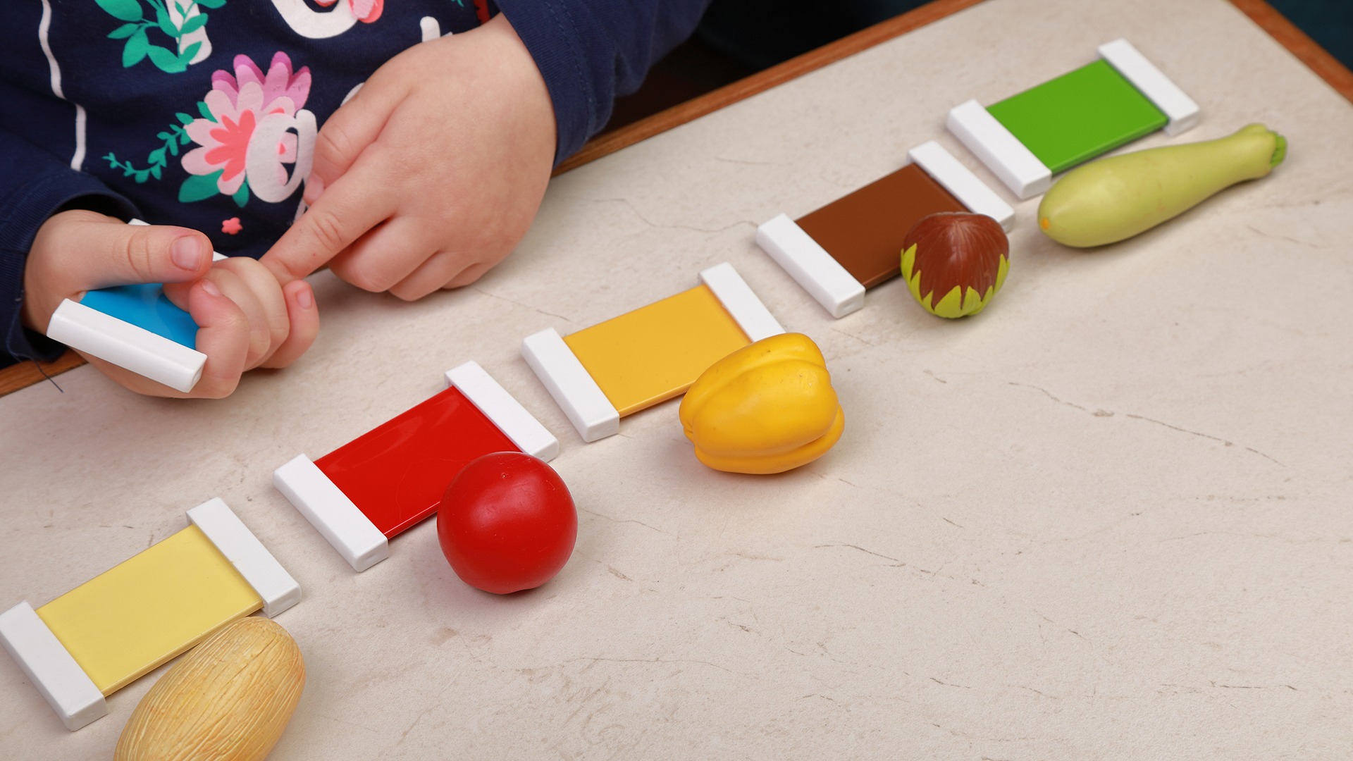 A child plays with colorful tiles and toy vegetables on a table, engaging in a creative sorting or matching activity.