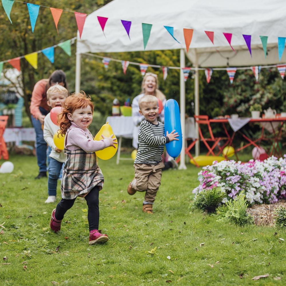 Children joyfully run with balloons in a garden, under a tent with colorful bunting. People and flowers surround the festive scene.