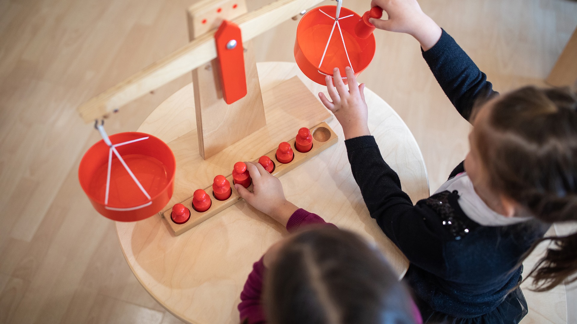 Two children playing with a wooden balance scale and red figurines on a round table, in a room with a light wooden floor.
