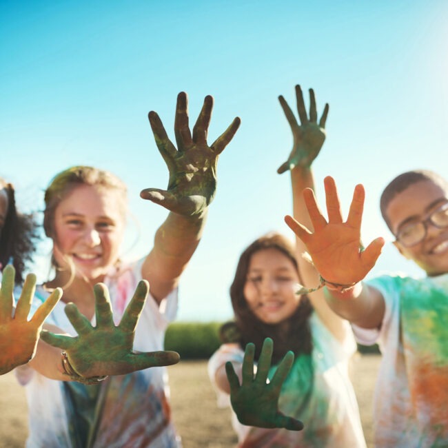 Four children smiling, reaching towards the camera with paint-covered hands, outdoors on a sunny day, blue sky in the background.