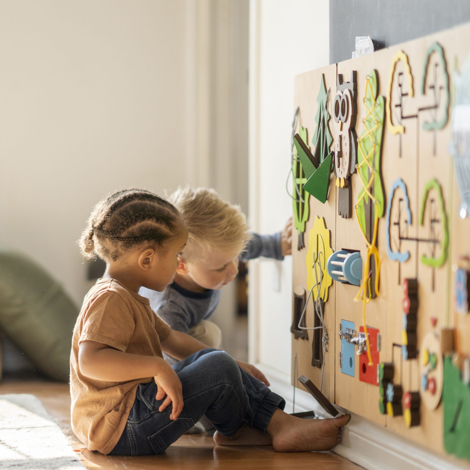Two children engaged with an interactive educational wall panel, exploring colorful shapes and activities, seated on a wooden floor in a bright room.