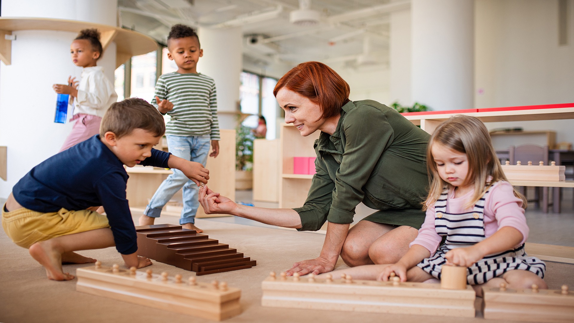 A person and four children engage in educational play with wooden toys, promoting learning and interaction in a bright, open classroom setting.