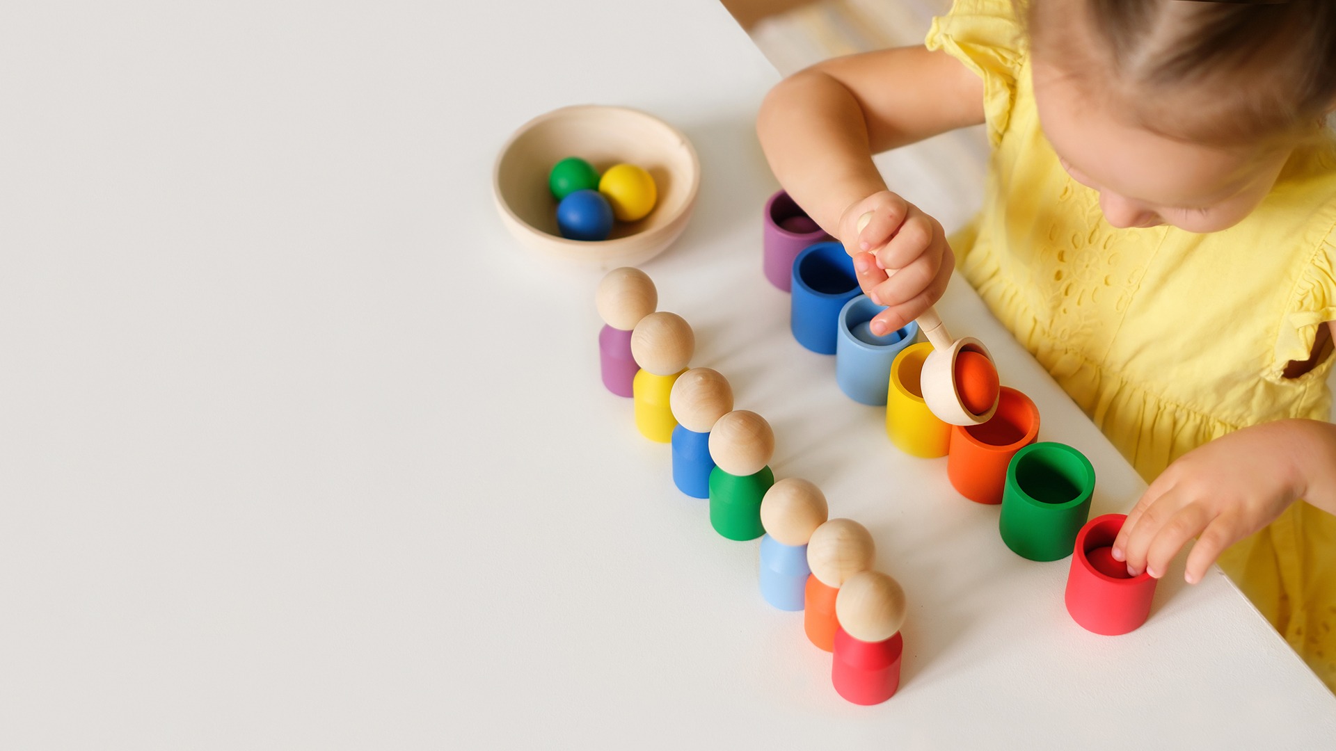 A child in a yellow dress plays with colorful wooden toys on a table, using a scoop to sort objects by color.