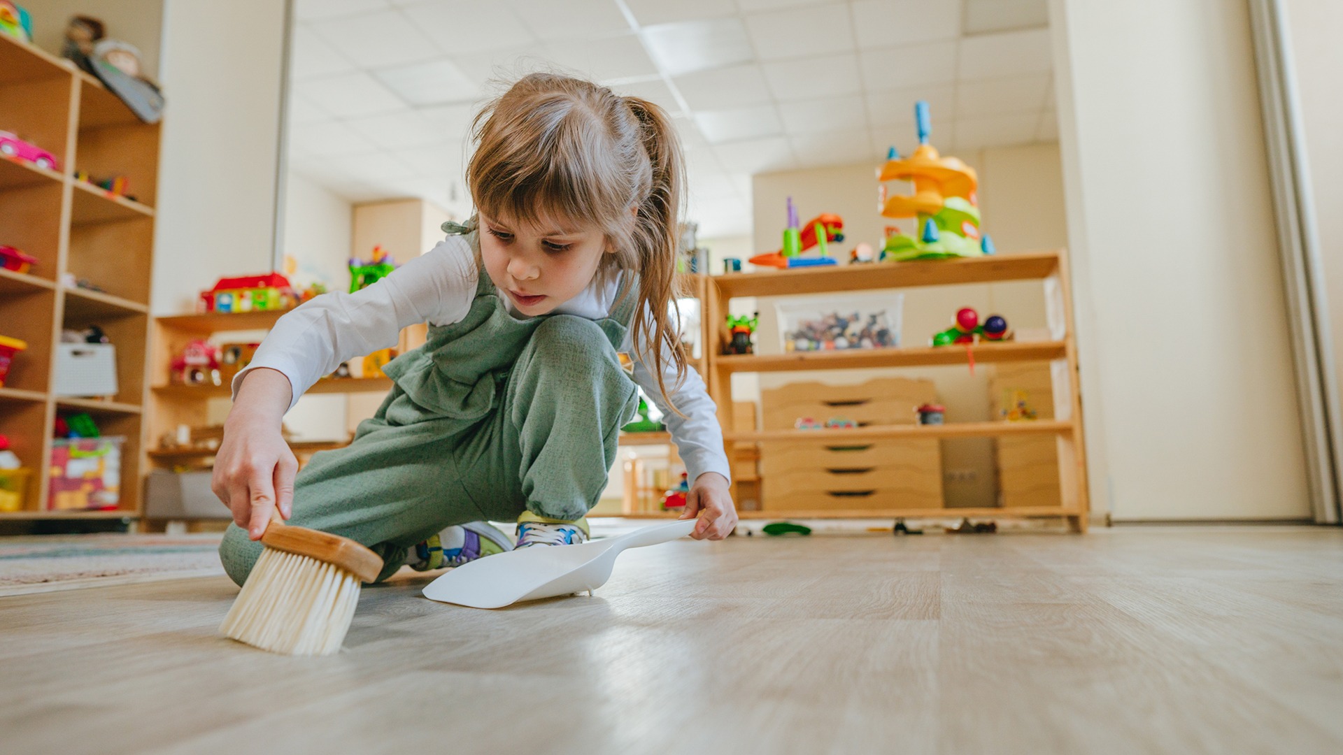 A child in overalls uses a brush and dustpan to clean a playroom with colorful toys and shelves in the background.
