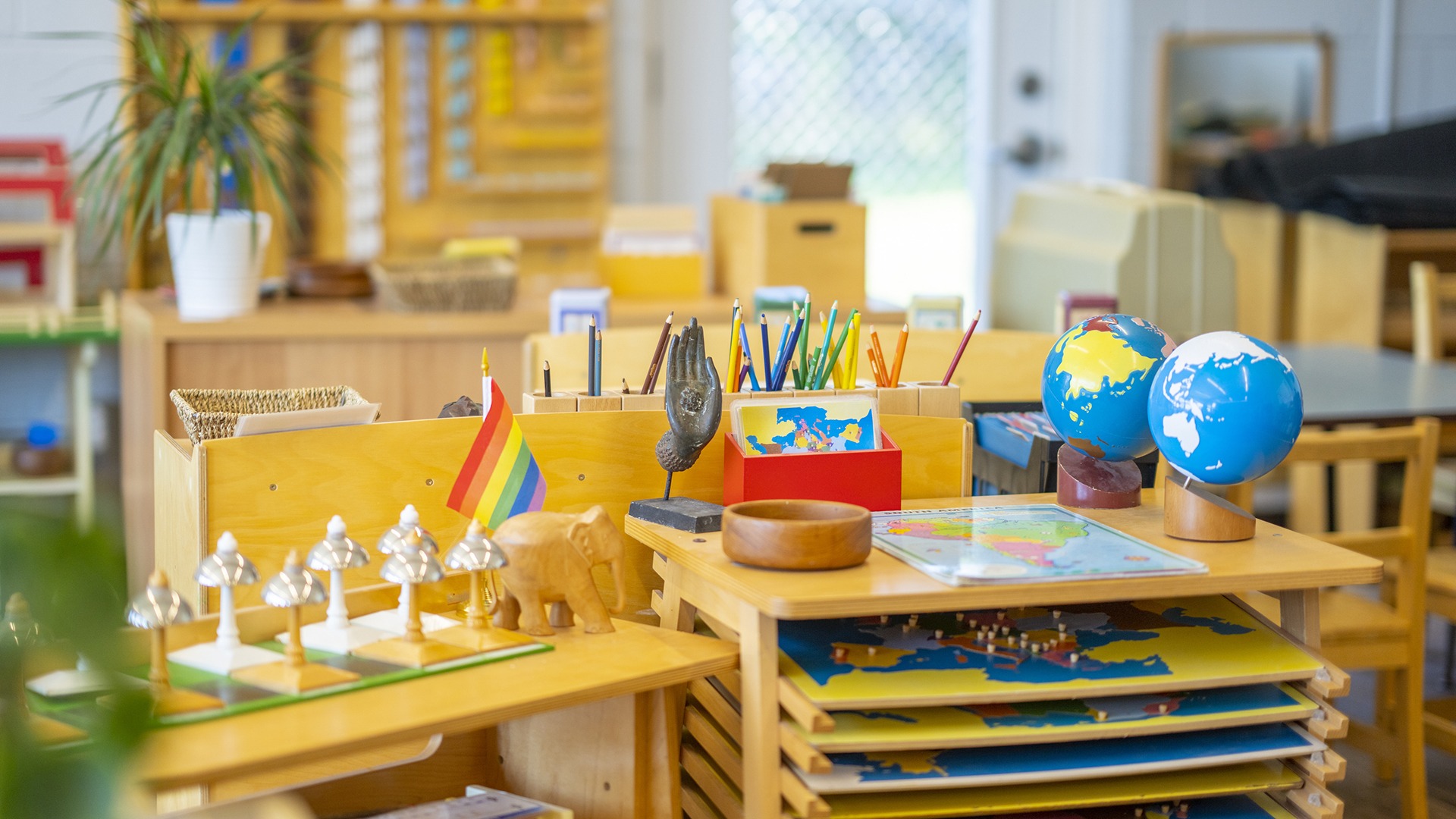 A colorful Montessori classroom features educational materials, including globes, a map, rainbow flag, and art supplies, promoting a diverse and interactive learning environment.