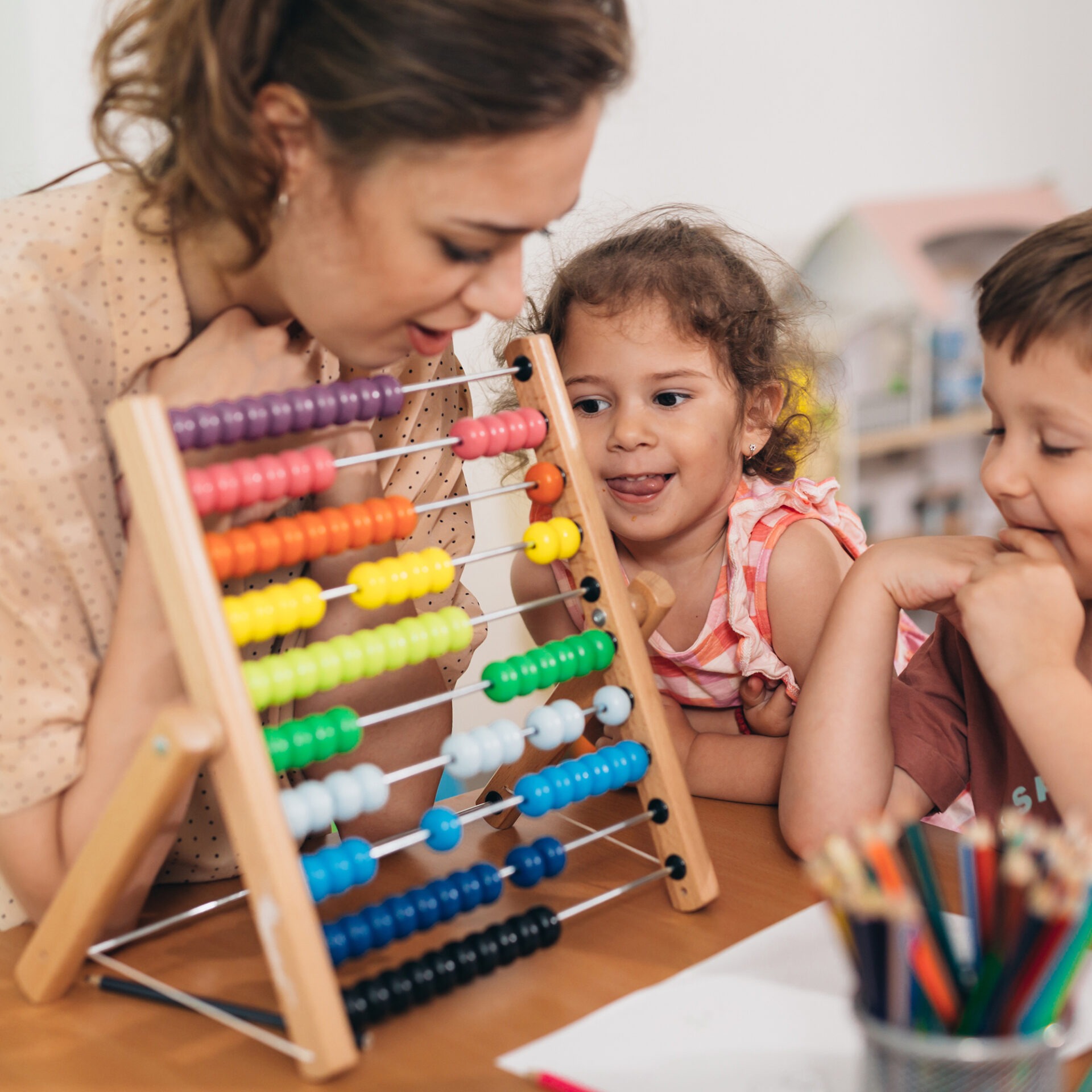 A person and two children engage with a colorful abacus at a table, surrounded by pencils and paper in a learning environment.