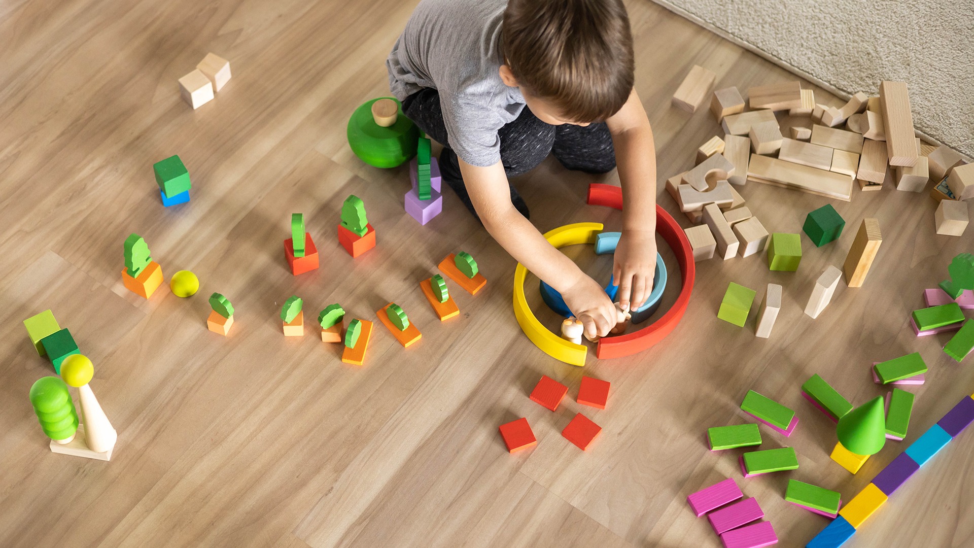 A child is playing with colorful wooden blocks on a wooden floor, arranging them into various shapes and structures, showing creativity.