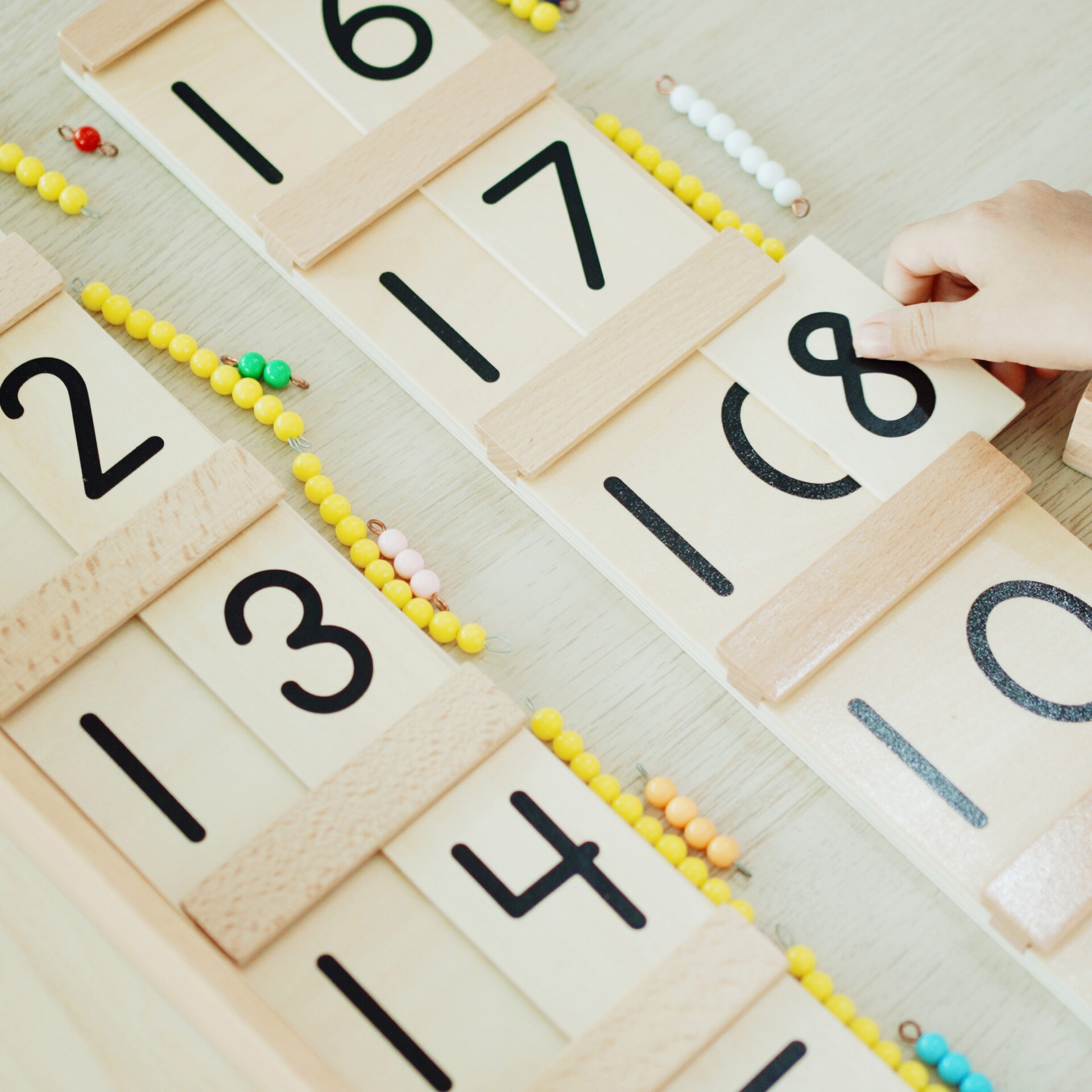 Wooden Montessori numbers with colorful bead strings are arranged on a table. A person's hand reaches for number eighteen.