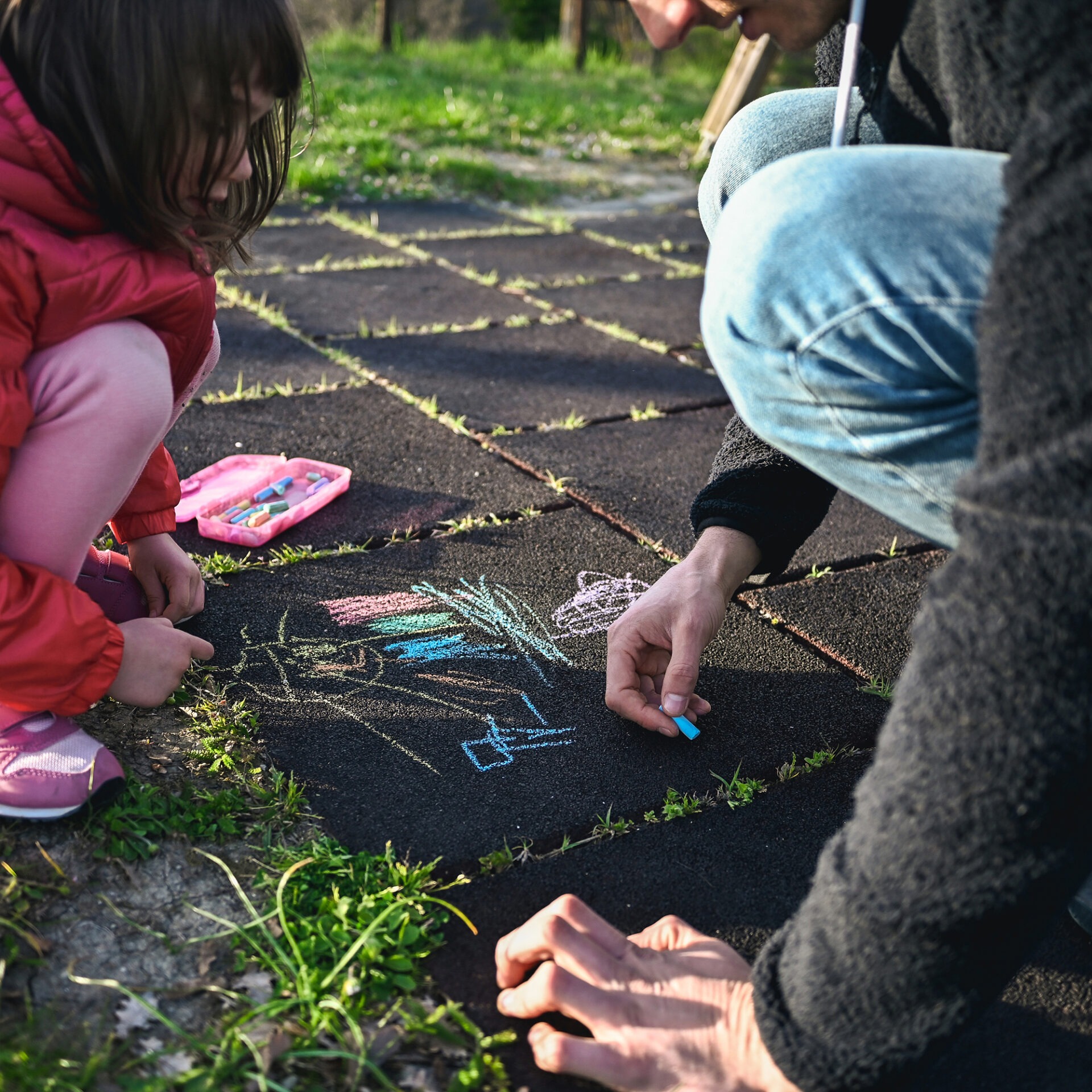 A young child and person draw colorful chalk pictures on pavement surrounded by grass on a sunny day.