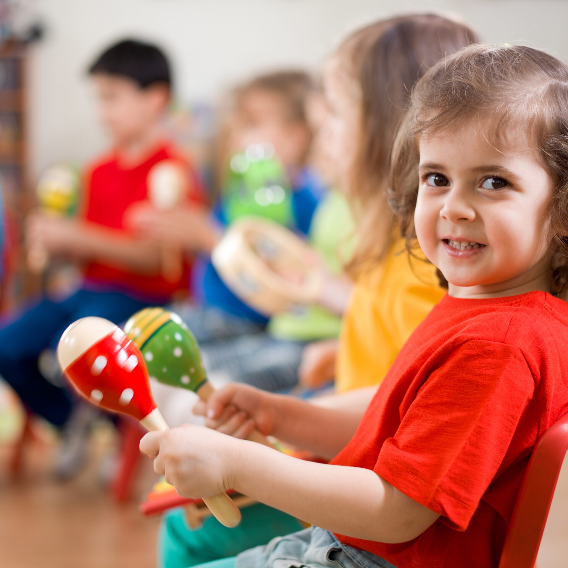 Children sitting in a row, playing musical instruments like maracas and tambourines, wearing colorful clothing, smiling and engaged in a lively activity.