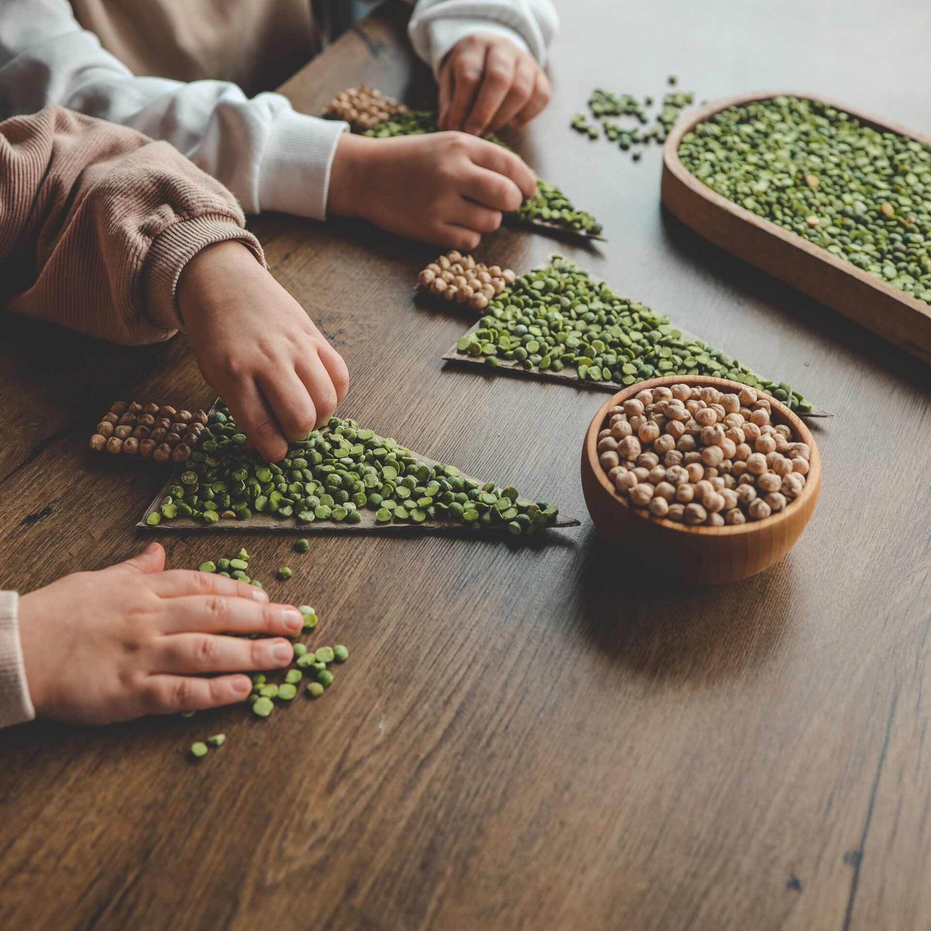 Hands arranging green and brown legumes on triangular pieces, with wooden bowls and beans on a wooden table in the background.