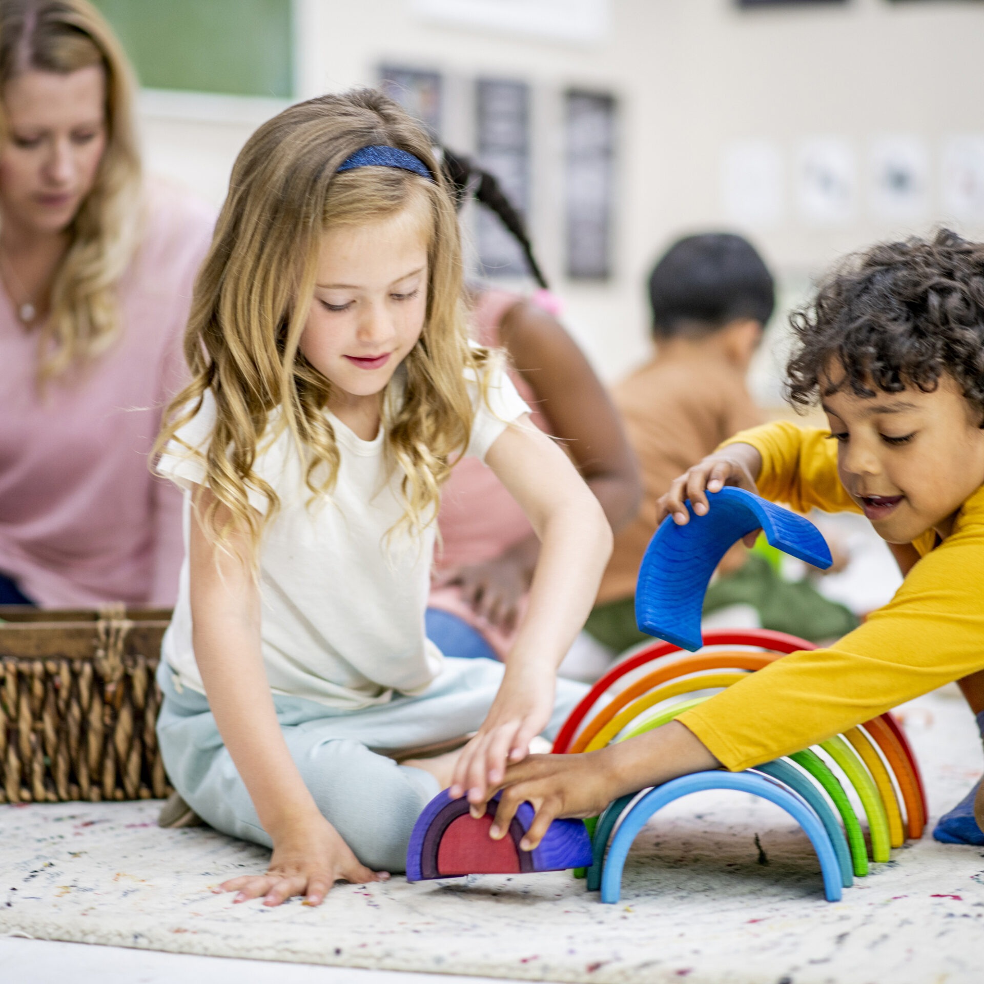 Children engage with colorful toy arches on a classroom floor, as a person watches nearby. Creativity and play in an educational setting.