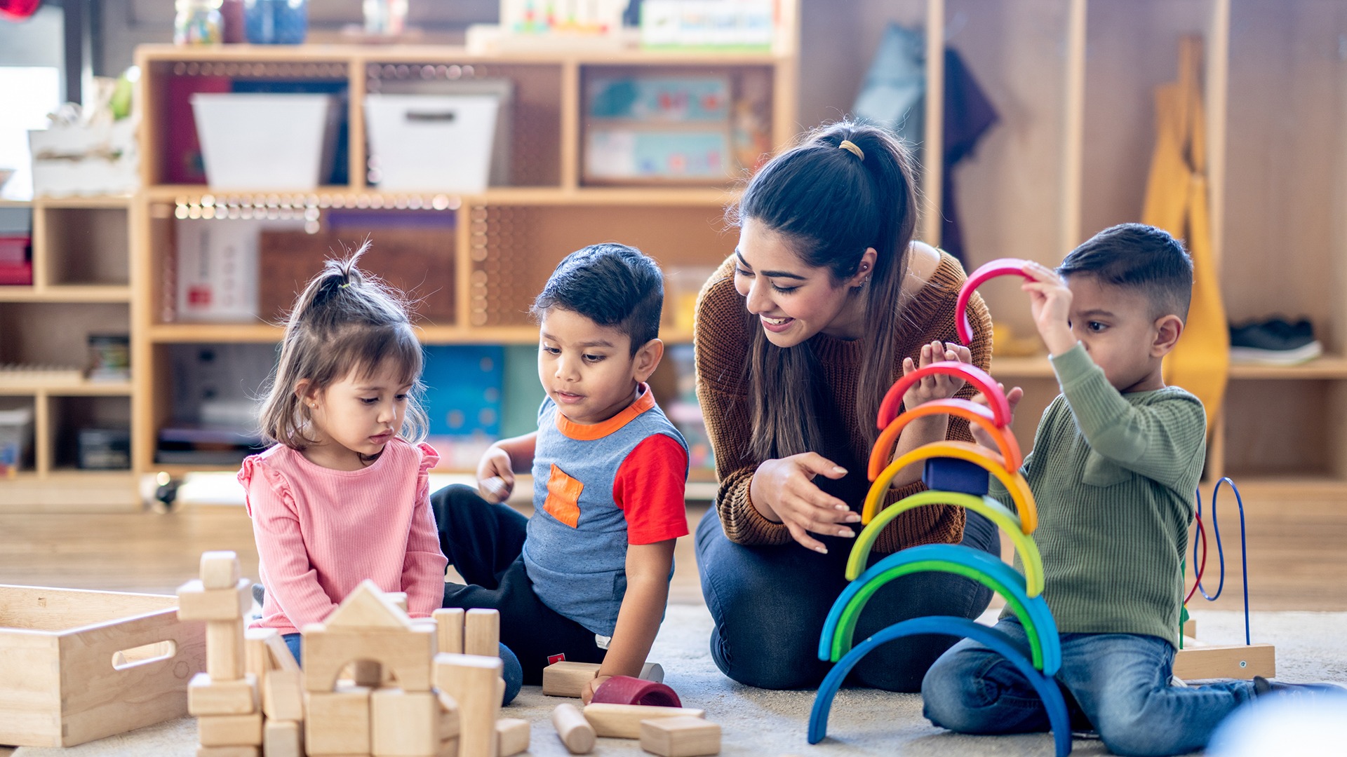 A person and three children engage in play with colorful toys and wooden blocks in a cozy, organized playroom.