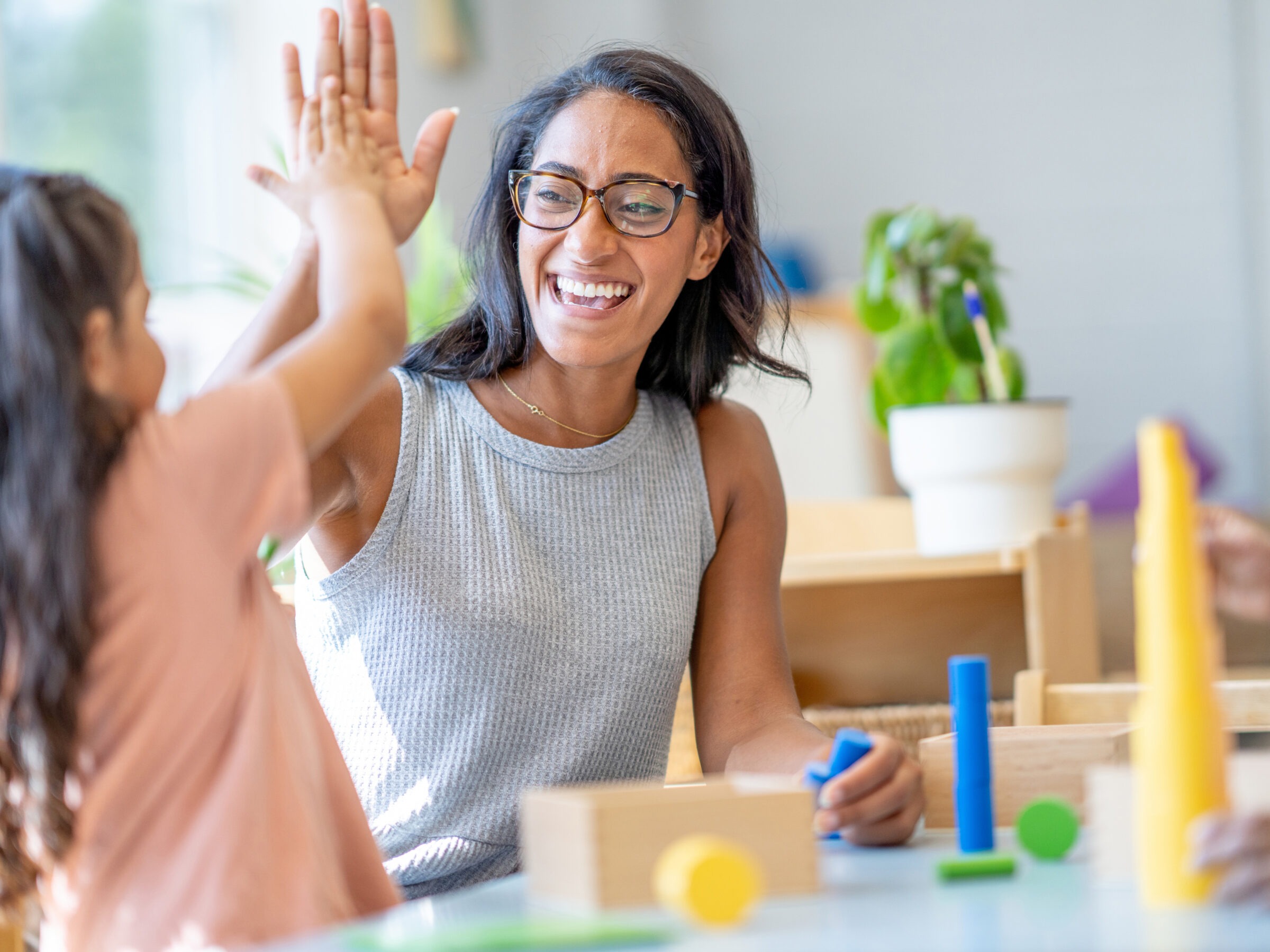 A person and child high-five in a bright room with toys and a plant, smiling and enjoying a playful moment.