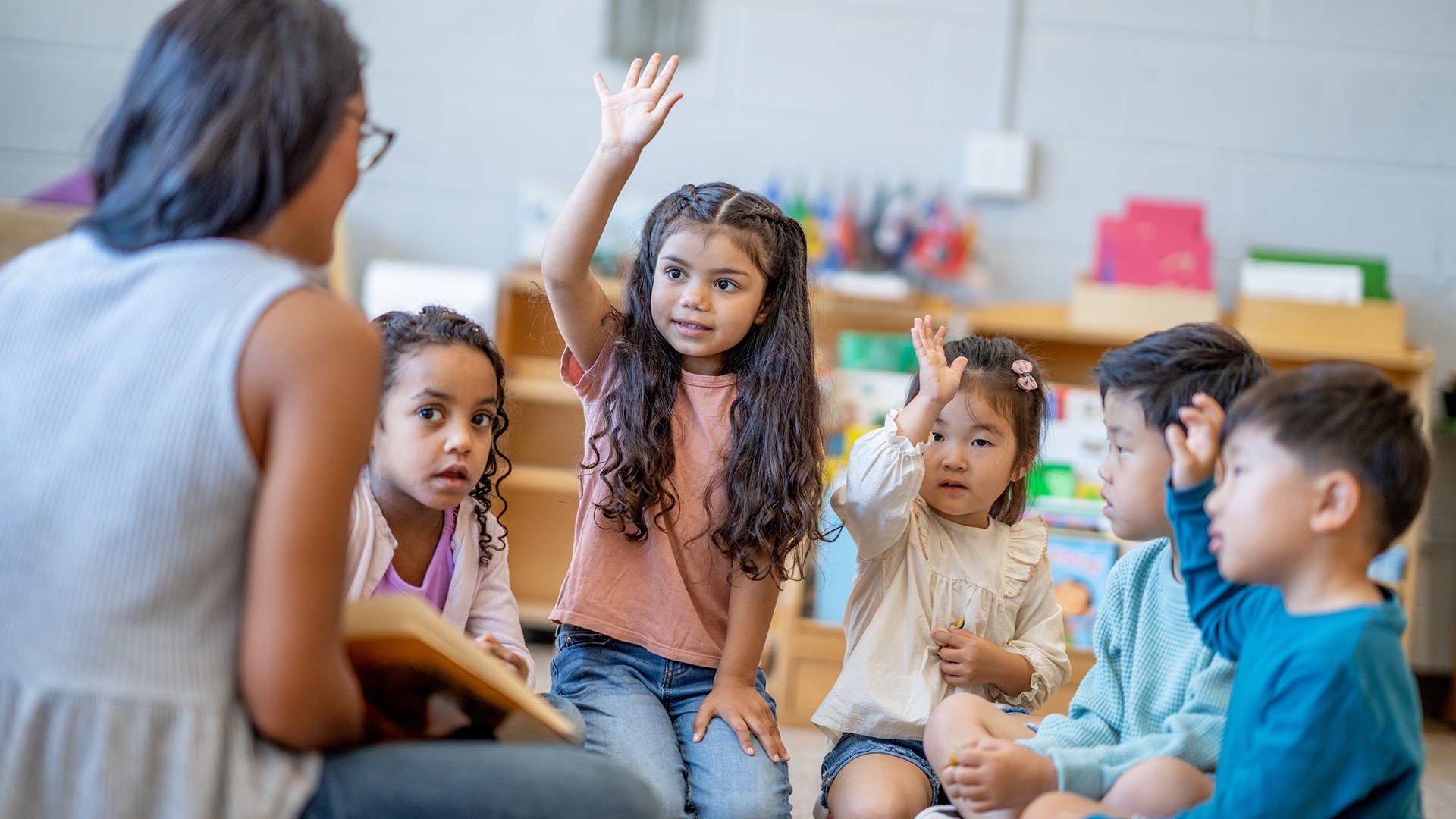 A person reads to attentive children in a classroom setting. Some children are raising their hands, engaging in the interactive learning session.