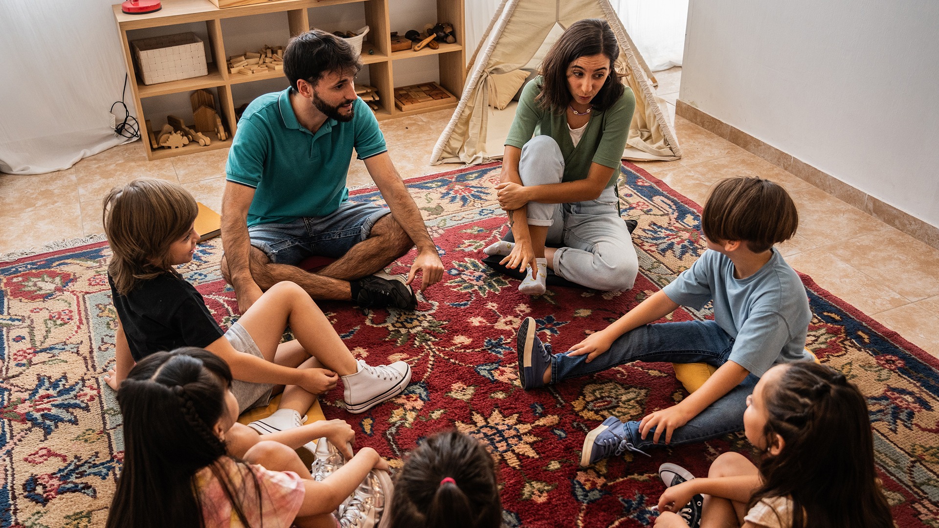 A group of children and two persons sit in a circle on a colorful rug, engaging in conversation in a cozy room.