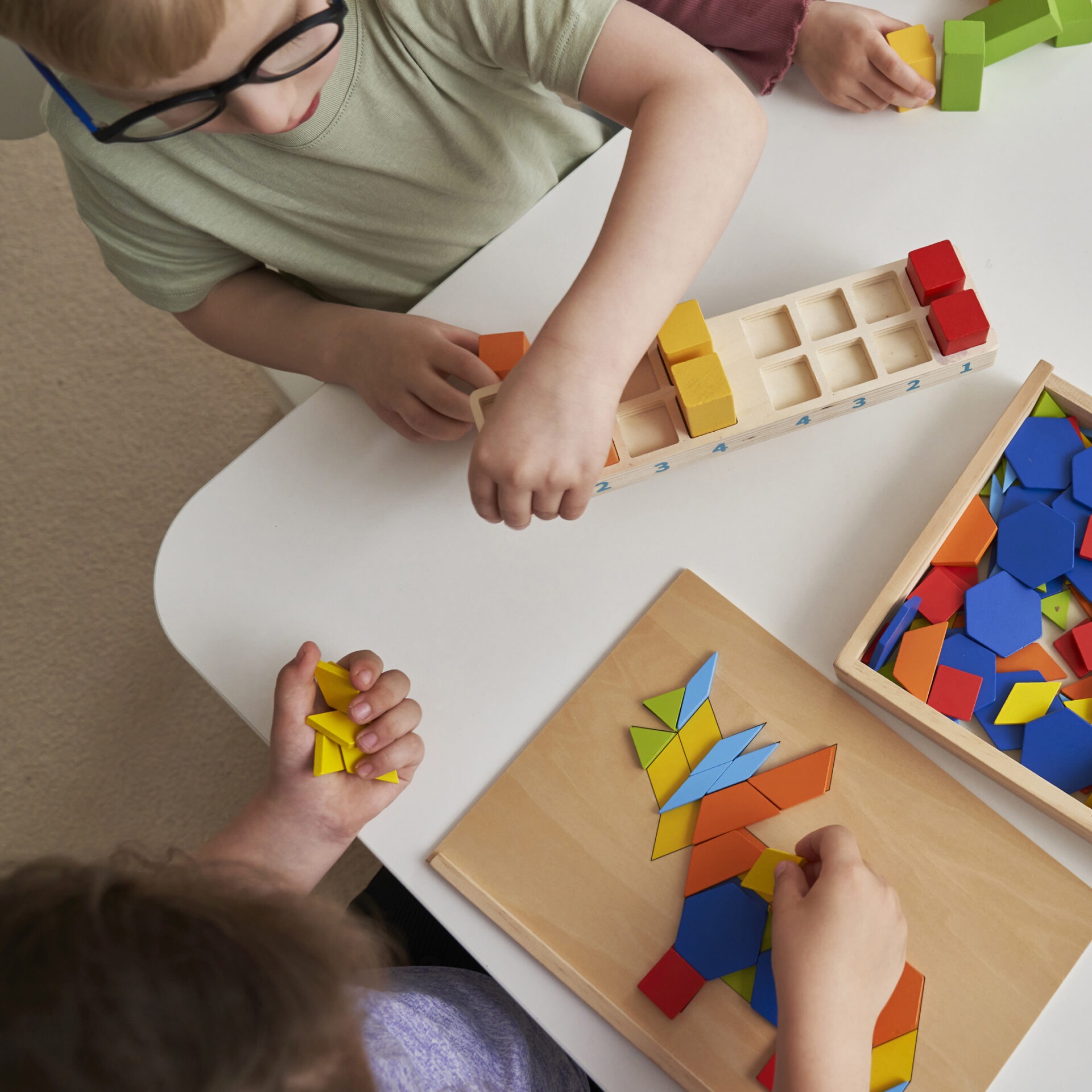 Children playing with colorful geometric shapes and blocks on a white table, engaging in creative and educational activities.