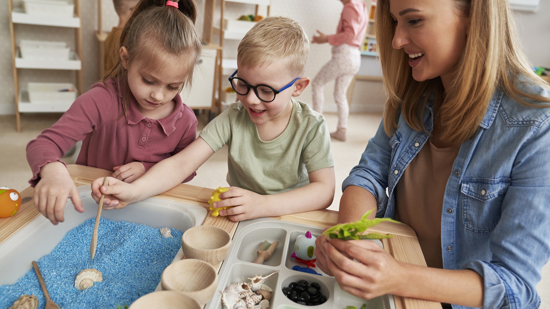 A person and two children engage in a sensory activity with blue rice and natural objects at a table in a classroom setting.