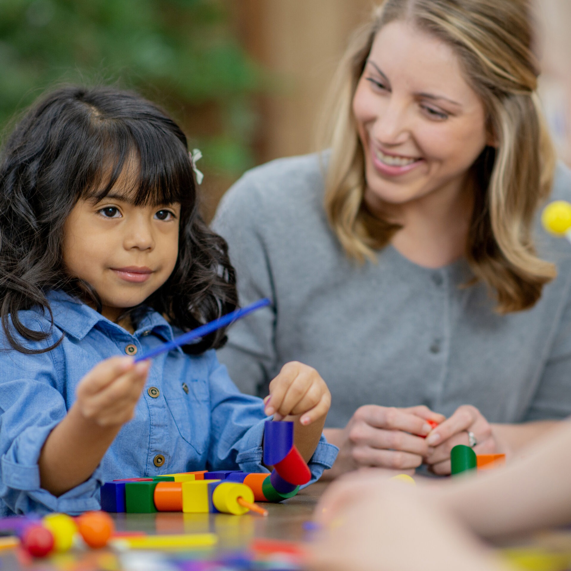 A child and person engage in a playful, creative activity with colorful building blocks and strings at a table, smiling in a cozy setting.