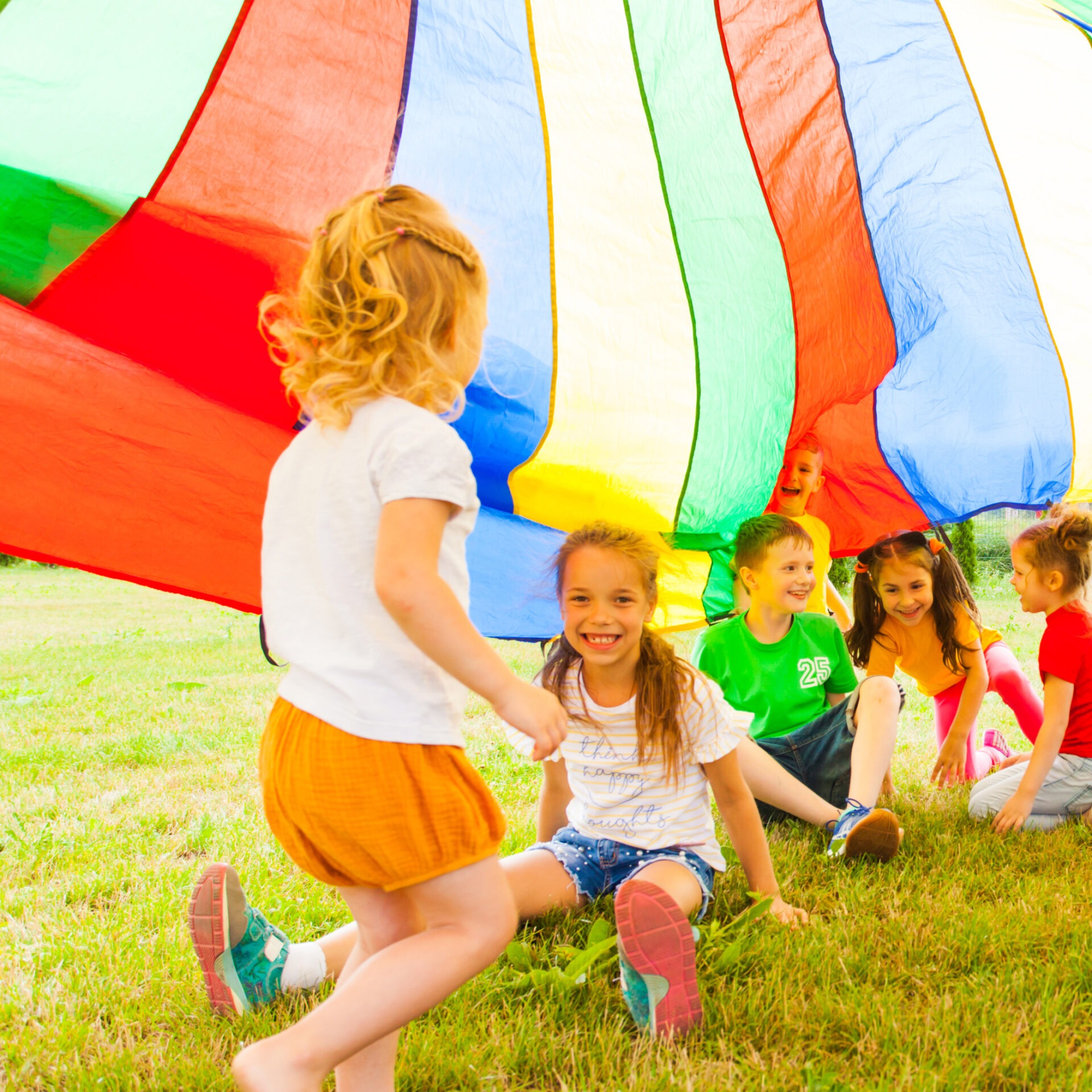 Children play under a colorful parachute tent on grassy field, smiling and interacting. Bright, cheerful atmosphere on a sunny day outdoors.
