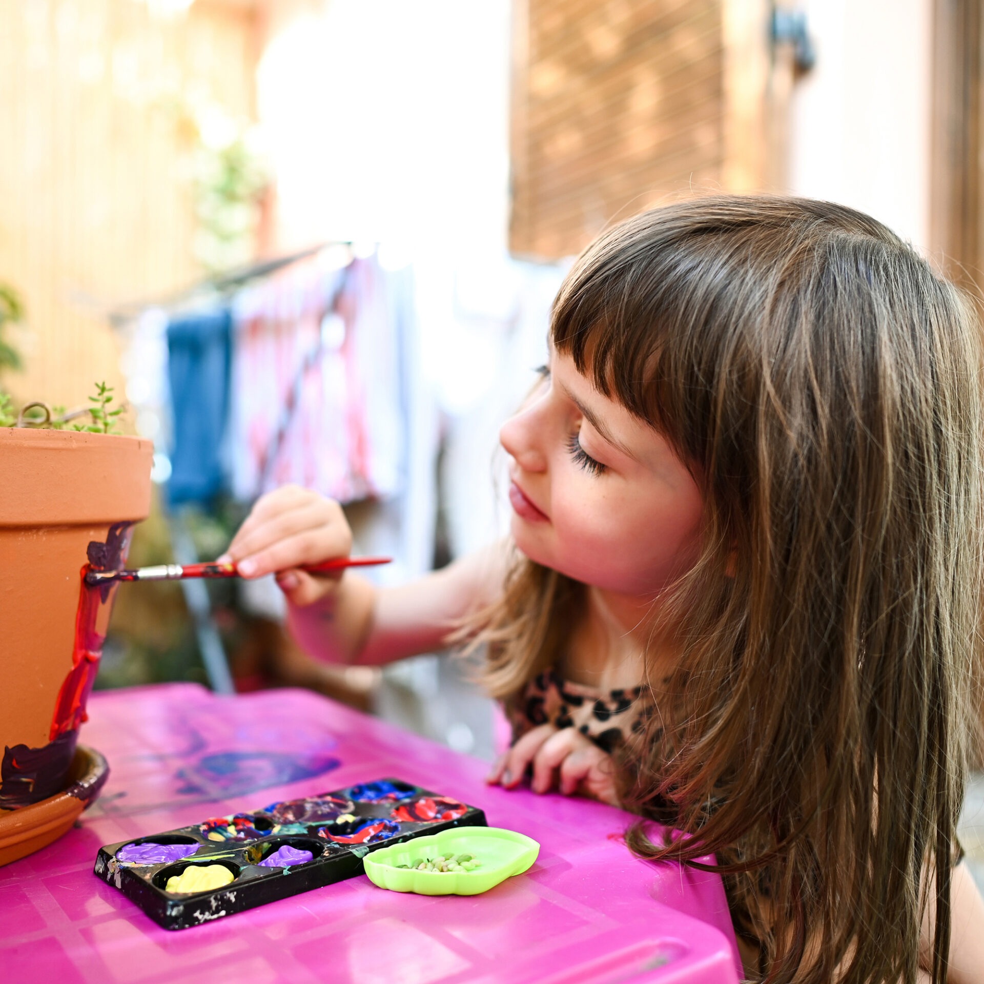 A child paints a terracotta pot with a brush, surrounded by colorful paint palette, engaged in a creative activity.