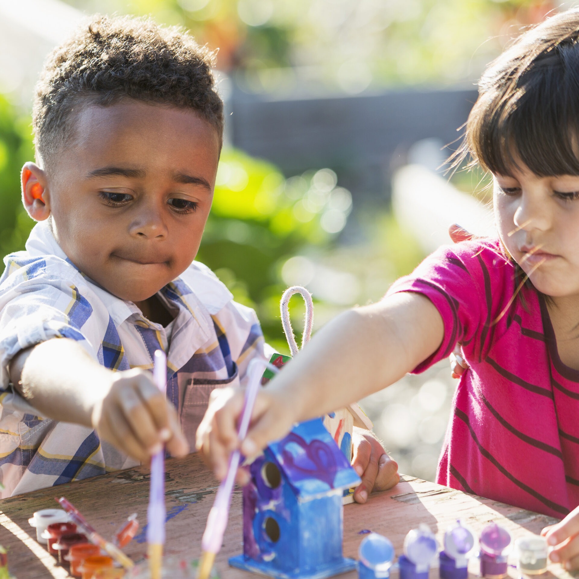 Two children are painting a small birdhouse outdoors, enjoying the creative activity with brushes and colorful paints on a sunny day.