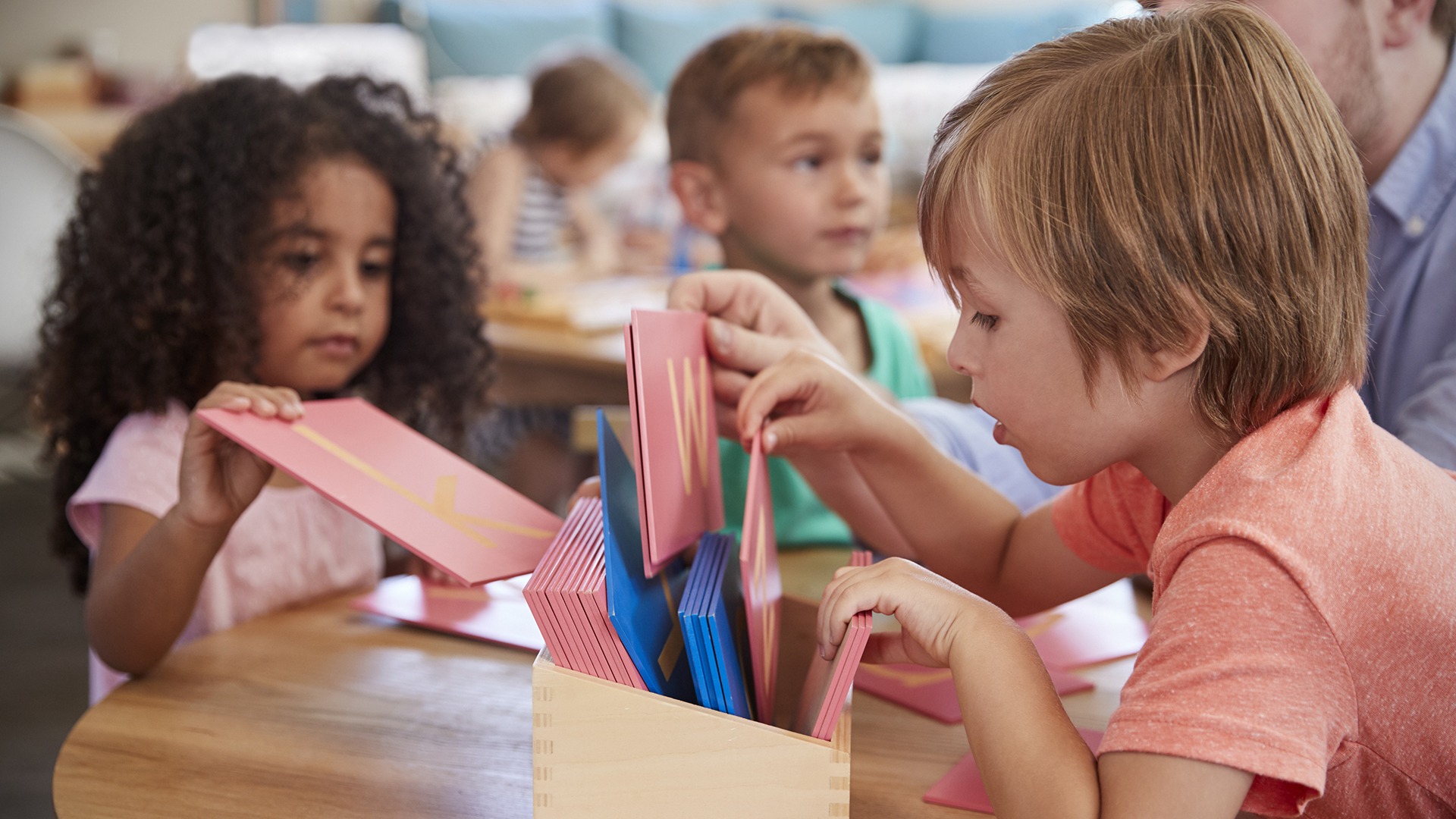 Children at a table engage with educational alphabet cards, focusing intently, while a person observes in a classroom setting.