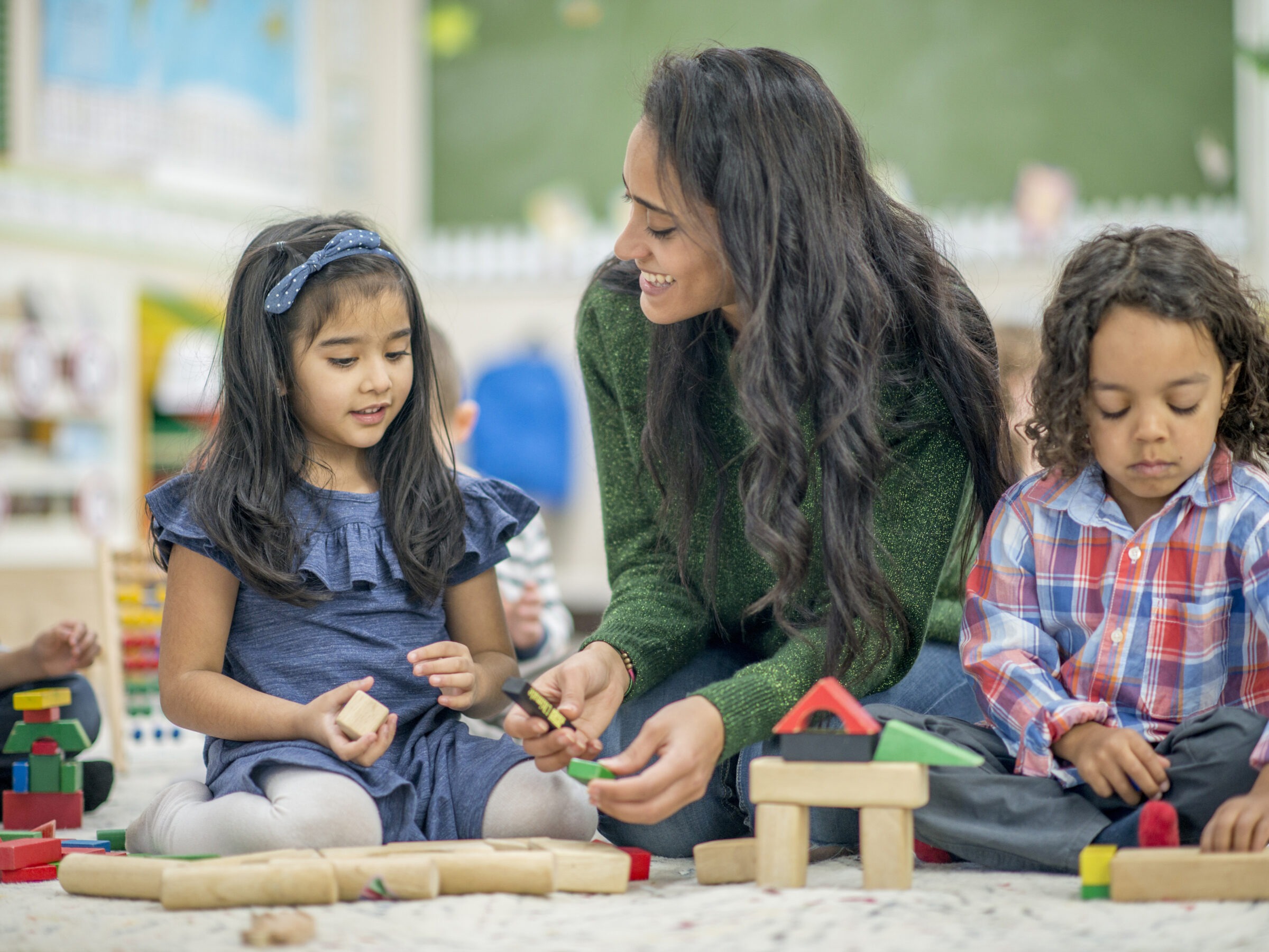 A person and two children are joyfully playing with building blocks in a colorful classroom, engaging in creative, educational activities together.