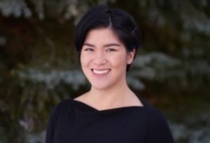 A person with short dark hair, wearing a black top, smiles against a blurred natural background of greenery. No landmarks or buildings visible.