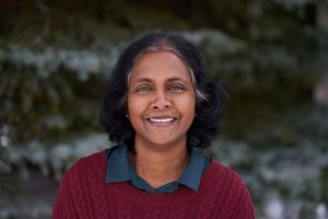 A person with short, dark hair smiles while wearing a red sweater over a collared shirt. The background features blurred greenery.