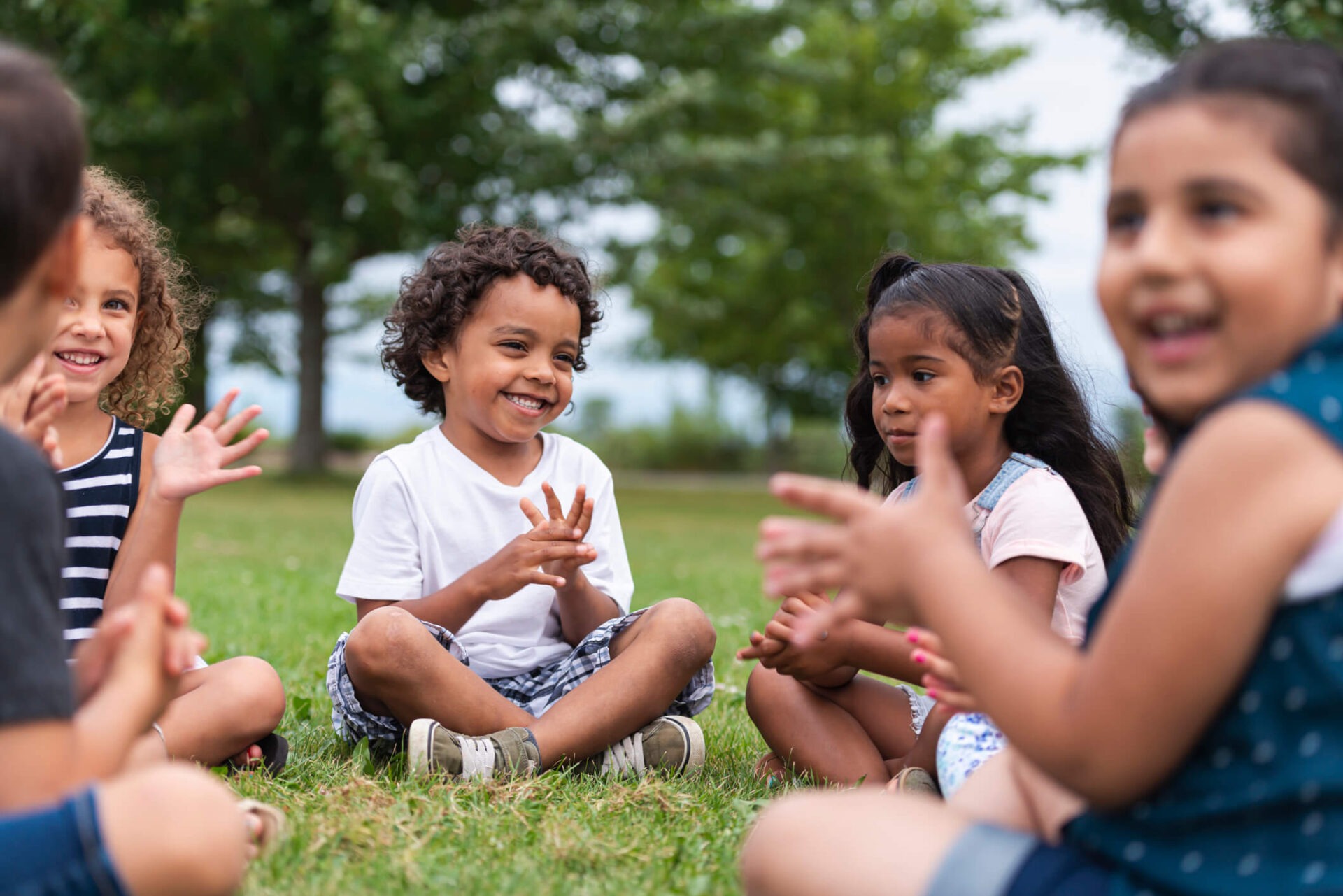 Children sitting on grass, smiling and clapping, surrounded by trees. Outdoor setting, bright atmosphere, group interaction, joyful expressions, casual clothing, and natural environment.