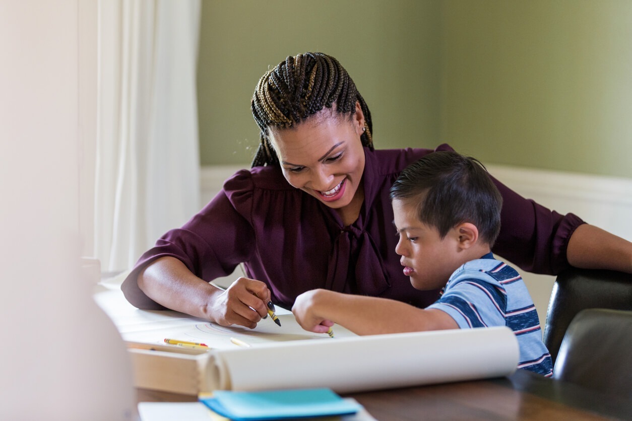 A person and child happily draw together at a table, surrounded by crayons and paper, in a warmly lit room.